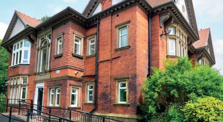 A red brick building with numerous windows, partially obscured by lush green bushes.