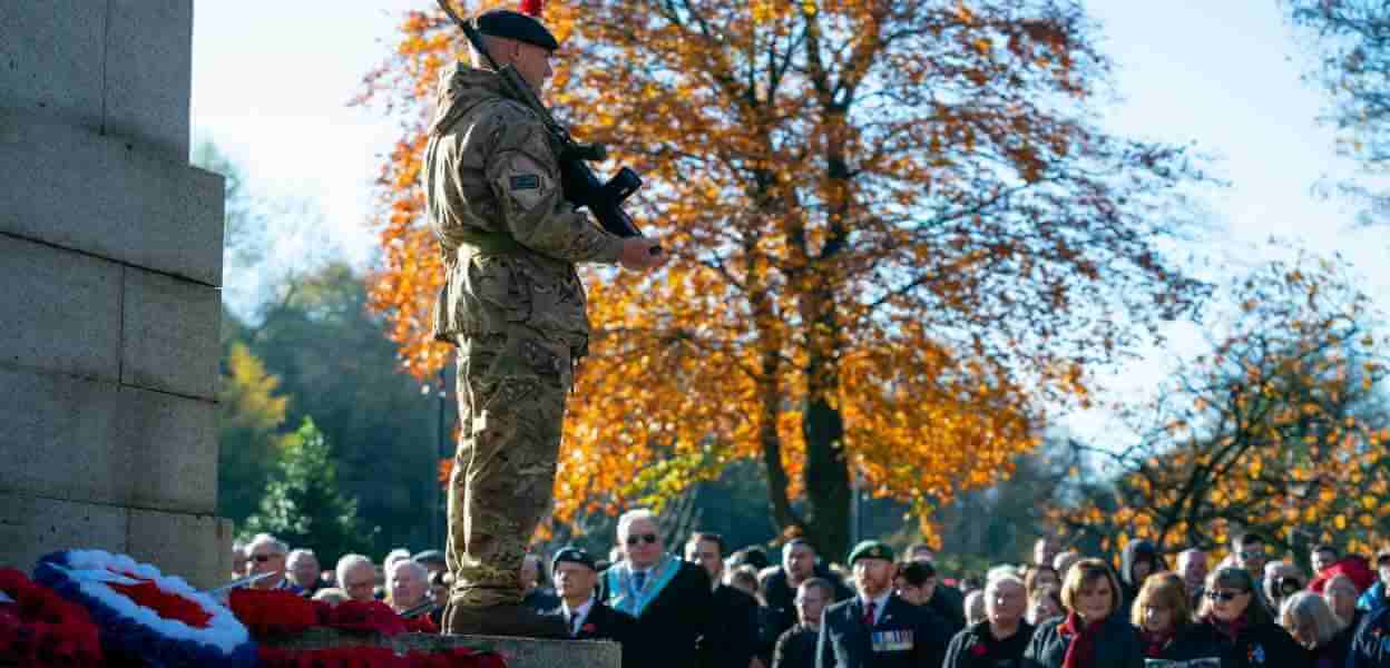 Veteran stood with rifle on steps of Rochdale Cenotaph with crowd gathered for Remembrance Sunday.