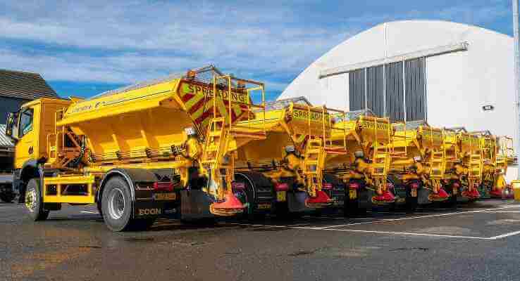 Row of parked gritters outside a salt storage shed.