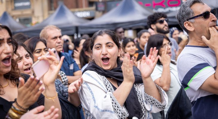 Crowd in a busy street scene with people clapping and cheering; a woman in the foreground smiles with hands raised.