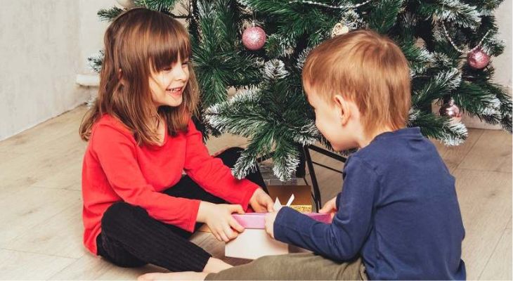 2 children opening presents by a Christmas tree.