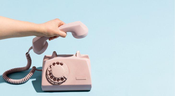 A hand lifting the receiver of a pink, retro rotary style phone.