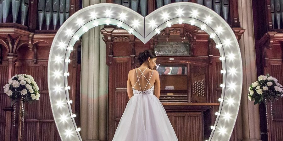 A woman in a wedding dress looking over her shoulder beneath an illuminated heart shaped arch.